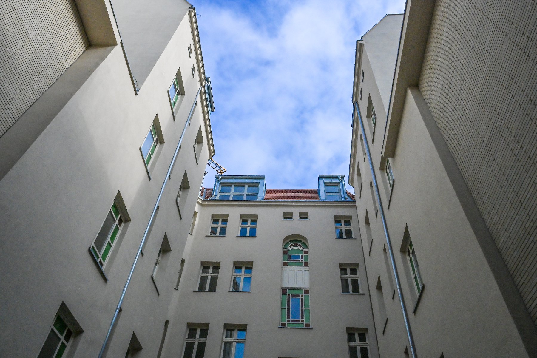 Perspektywiczne ujęcie z dołu przedstawiające wąski dziedziniec otoczony wysokimi, jasnymi ścianami kamienic. W centralnej części widać fasadę z ozdobnymi, witrażowymi oknami o zielonych i niebieskich szybkach. Powyżej znajduje się fragment dachu z czerwoną dachówką i lukarnami, a nad całością góruje błękitne niebo z delikatnymi chmurami.