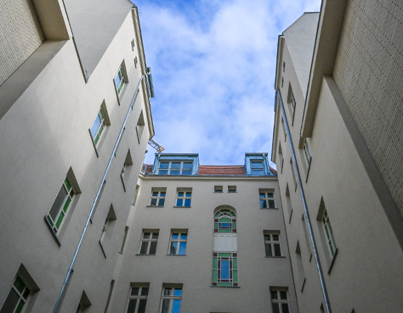 Perspektywiczne ujęcie z dołu przedstawiające wąski dziedziniec otoczony wysokimi, jasnymi ścianami kamienic. W centralnej części widać fasadę z ozdobnymi, witrażowymi oknami o zielonych i niebieskich szybkach. Powyżej znajduje się fragment dachu z czerwoną dachówką i lukarnami, a nad całością góruje błękitne niebo z delikatnymi chmurami.