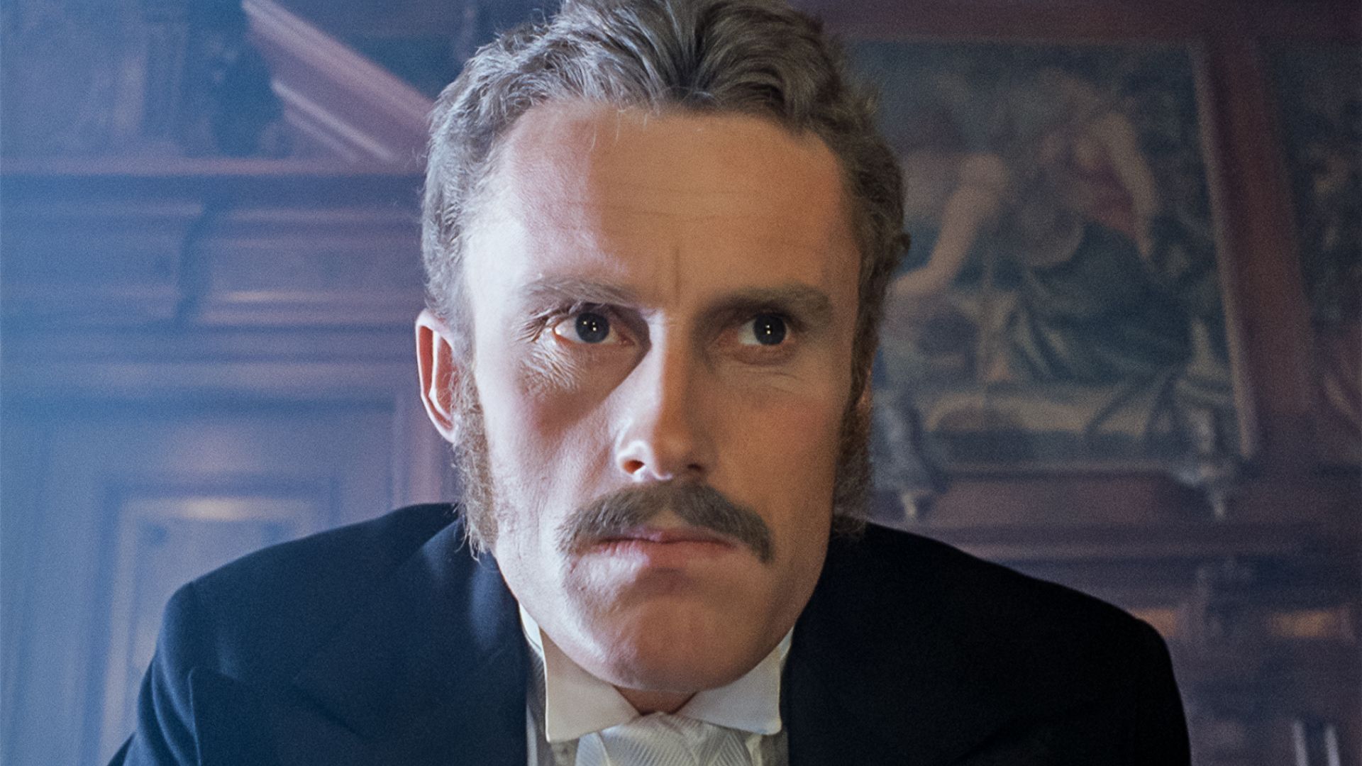 Portretowe ujęcie jasnowłosego mężczyzny z wąsami i baczkami, ubranego w ciemny garnitur z białą koszulą i muszką. Mężczyzna ma surowy wyraz twarzy i patrzy prosto przed siebie. W tle widoczne jest ciemne, drewniane wnętrze z fragmentem dużego obrazu na ścianie.