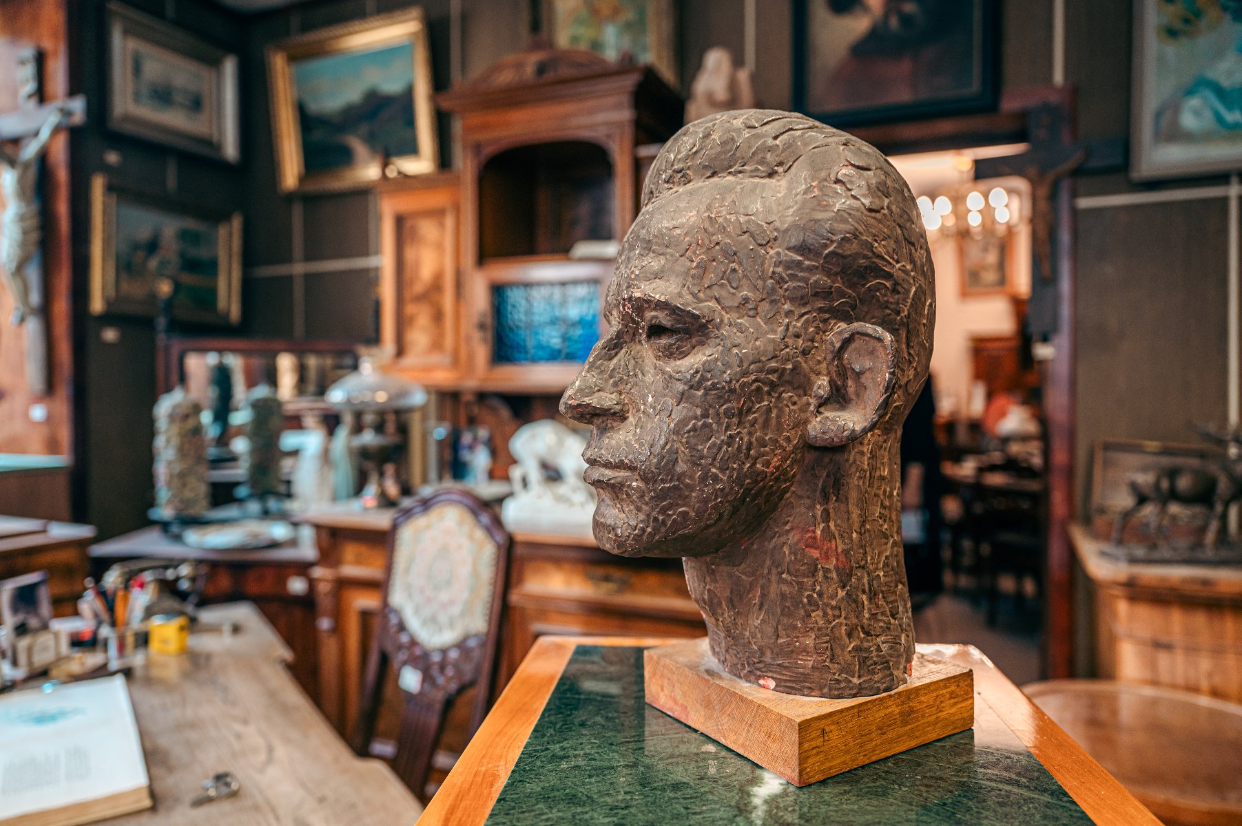 Rzeźba głowy mężczyzny wykonana z ciemnego, chropowatego materiału, ustawiona na drewnianej podstawie i zielonym postumencie. Popiersie ukazane jest z profilu. W tle widoczne jest bogato wyposażone wnętrze gabinetu lub antykwariatu: rzeźbione szafy, liczne obrazy w złotych ramach na ścianach, krzesło z ozdobną tapicerką oraz biurko z akcesoriami piśmienniczymi