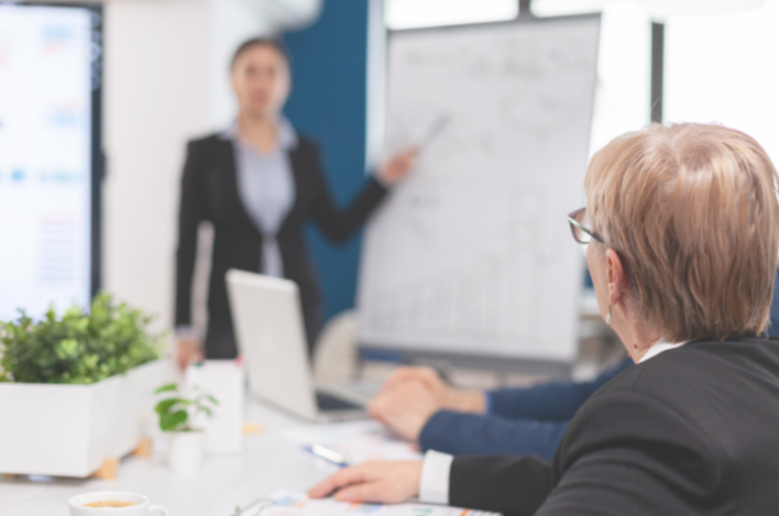 Poznań Biznes Partner zajmuje się organizacją i prowadzeniem różnorodnych programów wsparcia Spotkanie biznesowe w nowoczesnym biurze. Na pierwszym planie stół z laptopem, dokumentami, filiżanką kawy i małą rośliną. Po lewej i prawej stronie siedzą dwie osoby, a trzecia stoi i wskazuje na tablicę, na której znajdują się różne wykresy, schematy blokowe oraz diagramy.
