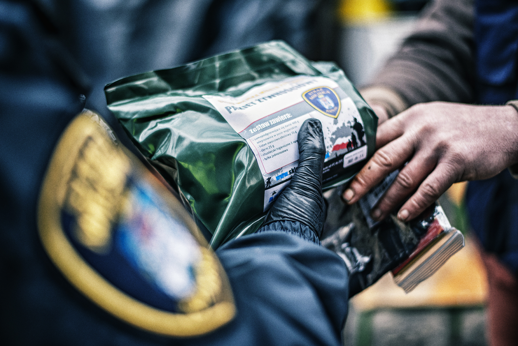 Ujęcie z bliska, gdzie widać dwie dłonie wymieniające paczkę. Ręka w czarnej rękawiczce i granatowym mankiecie kurtki trzyma zieloną, foliową paczkę, prawdopodobnie z racją żywnościową.