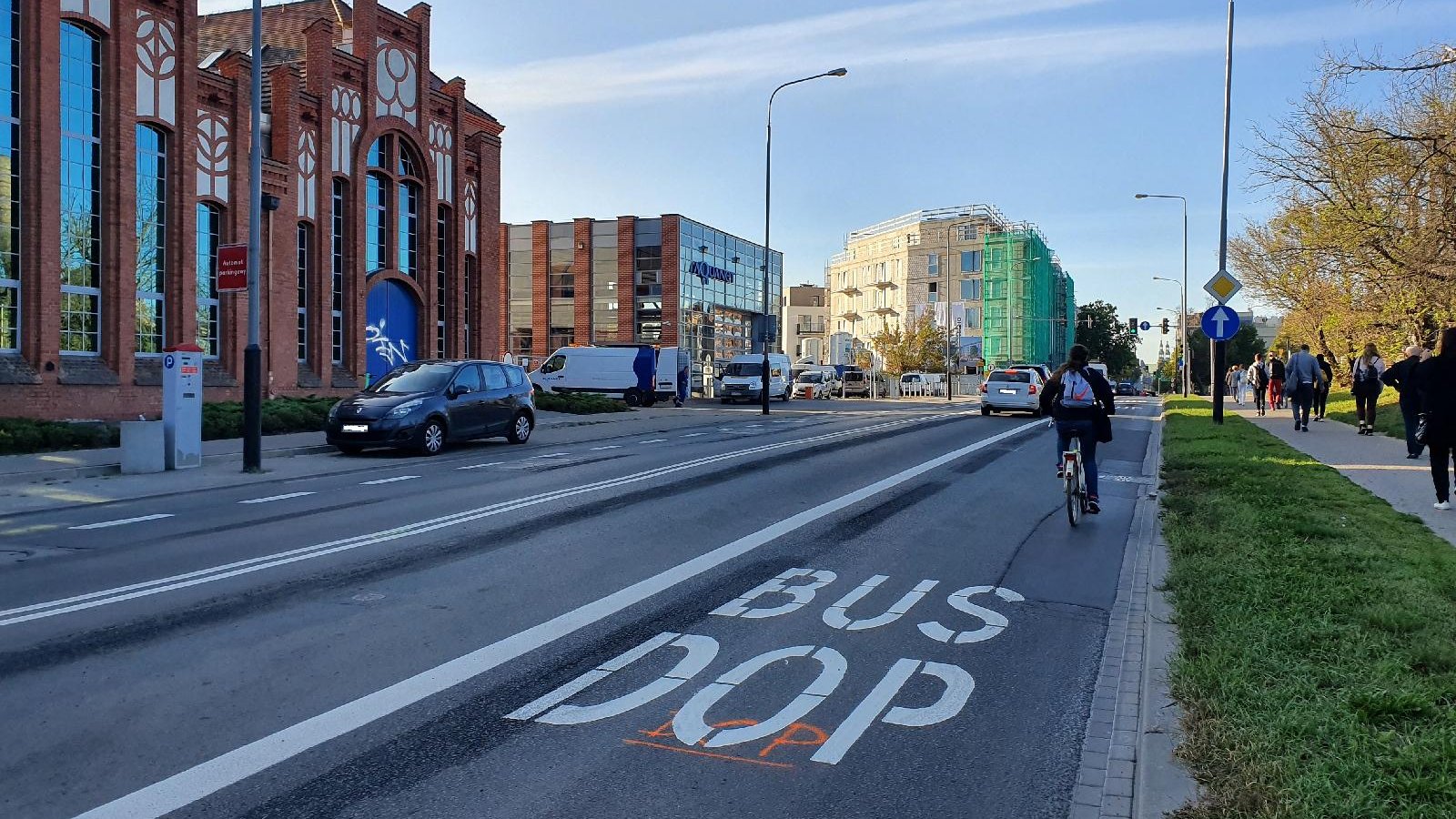 Ulica Garbary z trzema pasami ruchu. Rowerzysta jedzie po buspasie. Na chodniku idą ludzie. Po prawej stronie budynki Aquanetu, a w tle nowobudowany budynek - grafika artykułu