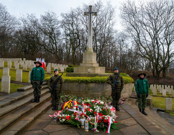 Uroczystość na cmentarzu wojennym. W centralnym punkcie znajduje się wysoki, kamienny krzyż z metalowym mieczem na postumencie, przed którym złożono liczne wieńce i zapalono znicze. Po obu stronach pomnika pełniona jest warta honorowa: stoją dwaj żołnierze w pełnym umundurowaniu z bronią oraz dwie harcerki w zielonych mundurach. W tle widoczne są rzędy jednakowych, jasnych tablic nagrobnych na trawiastym zboczu oraz bezlistne drzewa.