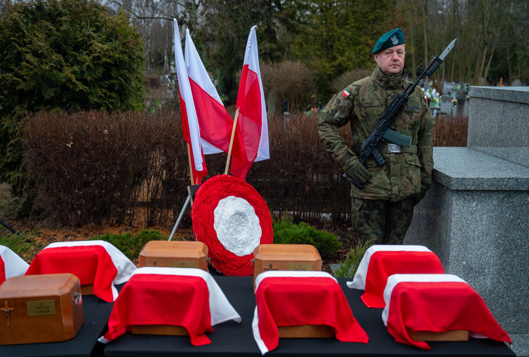 Uroczystość żałobna na cmentarzu. Na pierwszym planie, na stole przykrytym czarnym suknem, ustawiono drewniane urny, z których część przykryta jest biało-czerwonymi materiałami. W tle stoi żołnierz w pełnym umundurowaniu z karabinem, pełniąc wartę honorową. Obok niego znajdują się dwie polskie flagi oraz okrągły wieniec w narodowych barwach.