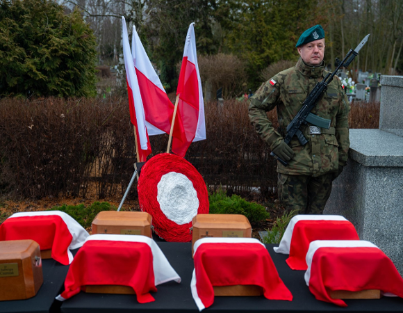 Uroczystość żałobna na cmentarzu. Na pierwszym planie, na stole przykrytym czarnym suknem, ustawiono drewniane urny, z których część przykryta jest biało-czerwonymi materiałami. W tle stoi żołnierz w pełnym umundurowaniu z karabinem, pełniąc wartę honorową. Obok niego znajdują się dwie polskie flagi oraz okrągły wieniec w narodowych barwach.