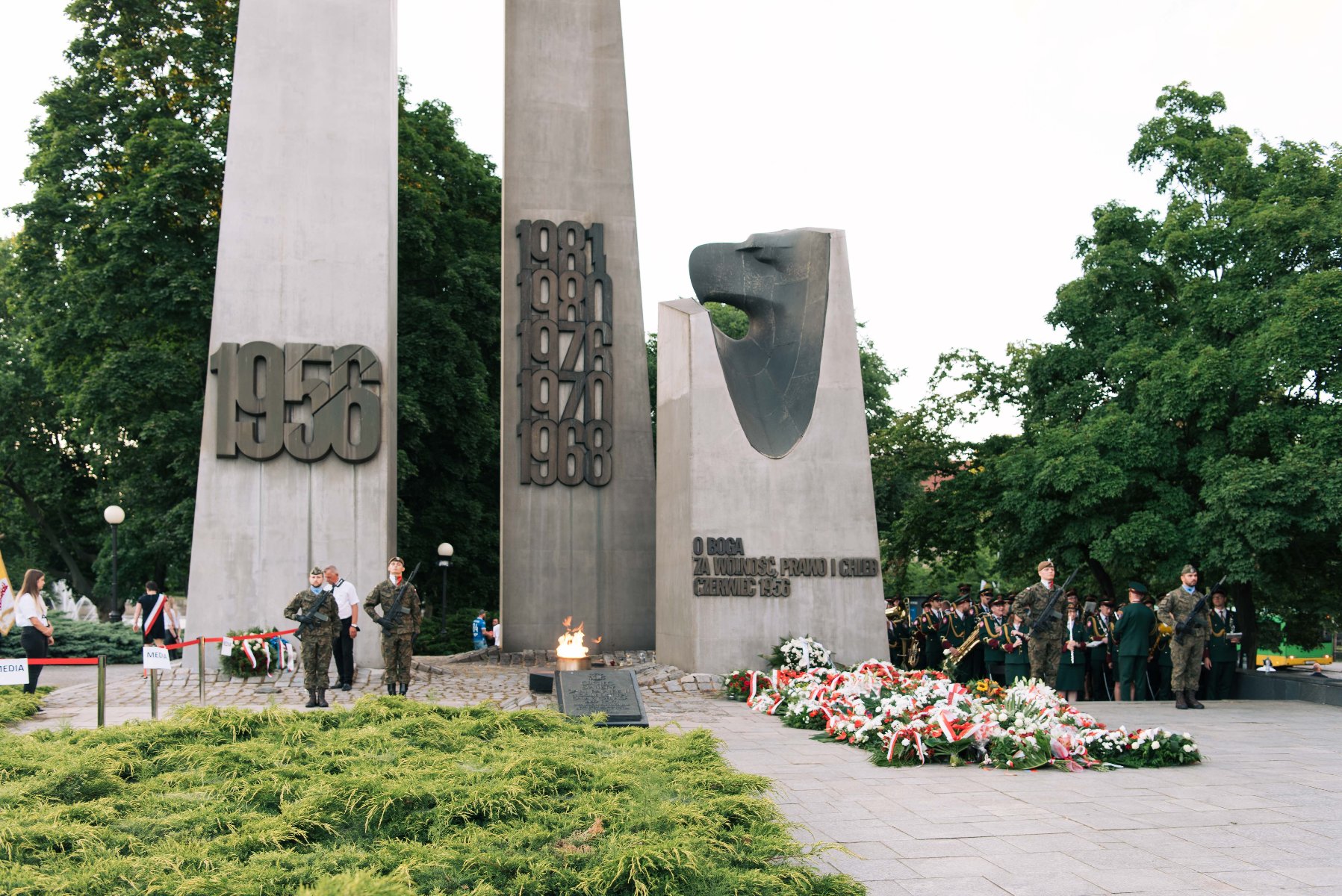 Uroczystości pod Pomnikiem Poznańskiego Czerwca 1956 na placu Mickiewicza. Widoczne są dwa wysokie, betonowe krzyże z datami 1956 oraz 1968, 1970, 1976, 1980 i 1981. Przed pomnikiem płonie znicz, a na ziemi ułożono liczne wiązanki biało-czerwonych kwiatów.
