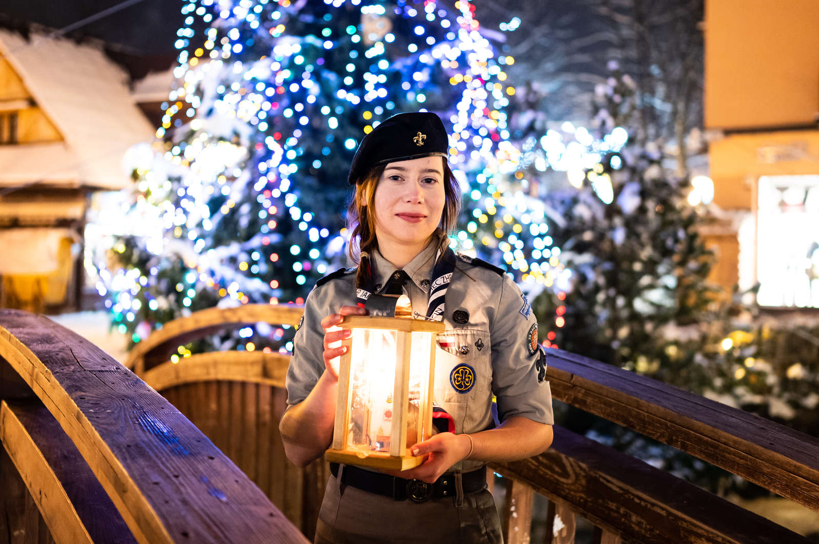 Uśmiechnięta młoda harcerka w szarym mundurze, ciemnym berecie i z odznakami, stoi na drewnianym, ośnieżonym mostku i trzyma w dłoniach lampion ze Światłem Betlejemskim. W tle widoczna jest duża, udekorowana choinka świąteczna z licznymi niebieskimi i białymi lampkami.