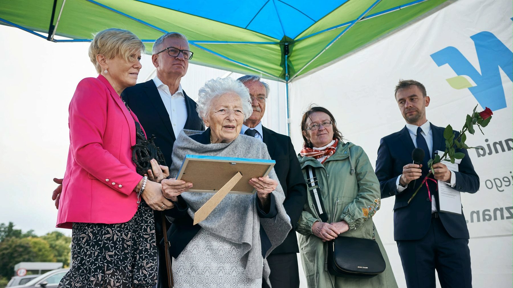 Na zdjęciu prezydent Poznania z seniorami, mężczyzna trzyma mikrofon i różę, kobieta trzyma ramkę ze zdjęciem