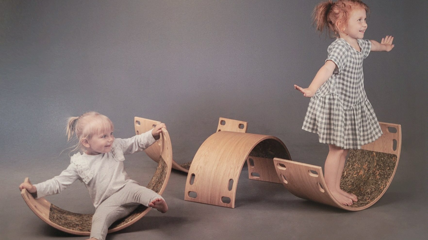 Na zdjęciu dwójka dzieci bawiąca się na drewnianych półokrągłych bujaczkach