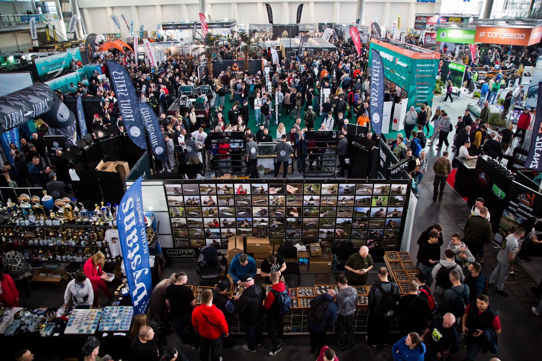 Widok z góry na zatłoczoną halę targową z licznymi stoiskami wędkarskimi. Na pierwszym planie stoisko z rzędami niewielkich akcesoriów w gablotach, przy którym gromadzą się ludzie. W tle widoczne wysokie banery reklamowe, między innymi z napisami "Dystans", "LIBRA LURES" oraz "torqeedo". W głębi hali tłum zwiedzających przemieszczający się między ekspozycjami.