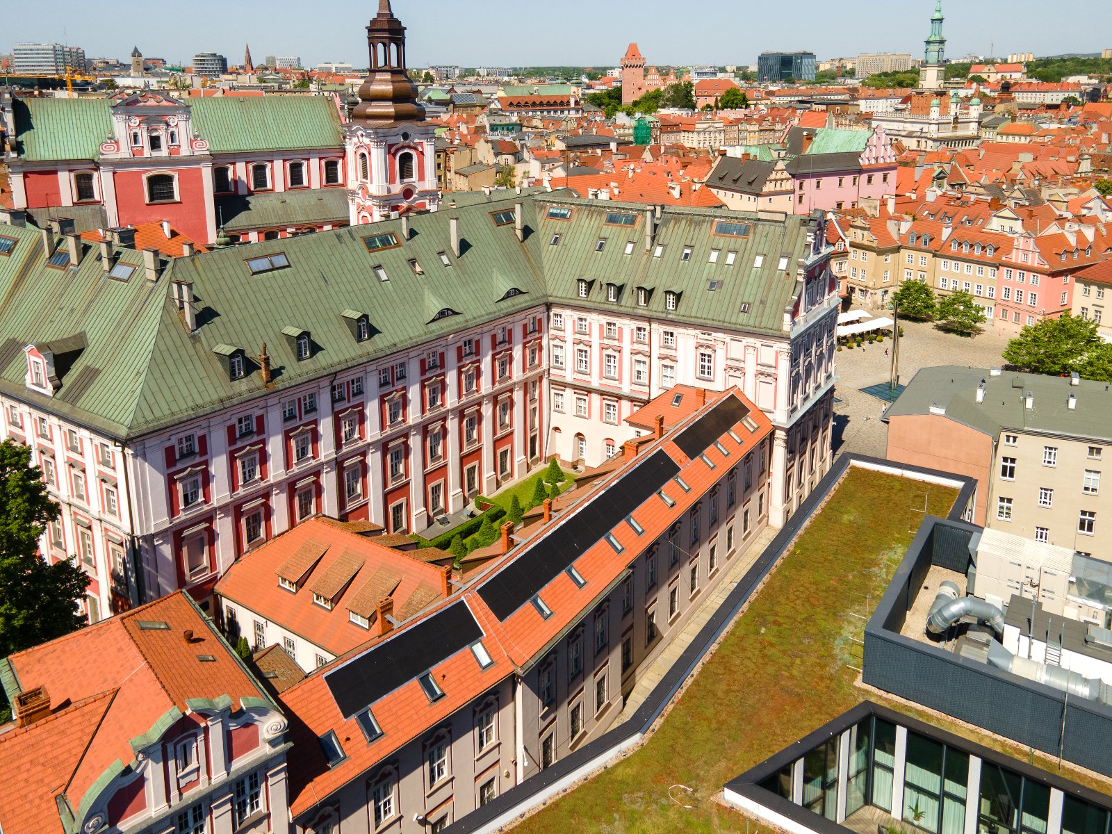 W piątek, 2 maja Urząd Miasta Poznania zapewni obsługę we wszystkich obszarach swojej działalności Widok z lotu ptaka na miasto Poznań. Centralny punkt zdjęcia to duży, zdobiony budynek z zielonym dachem i czerwonymi akcentami, otoczony budynkami z czerwonymi dachówkami. W tle widać wieże i budowle reprezentujące różnorodne style architektoniczne.