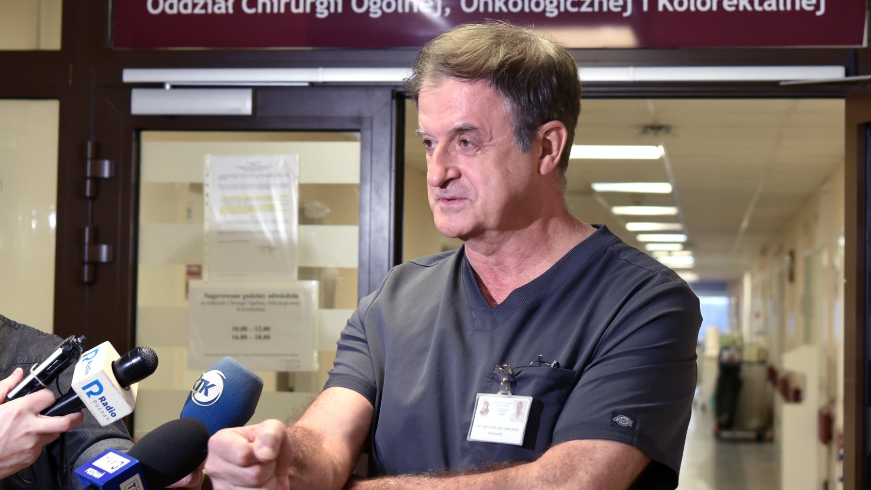 Na zdjęciu chirurg mówiący coś do mikrofonu, stojący w korytarzu szpitala