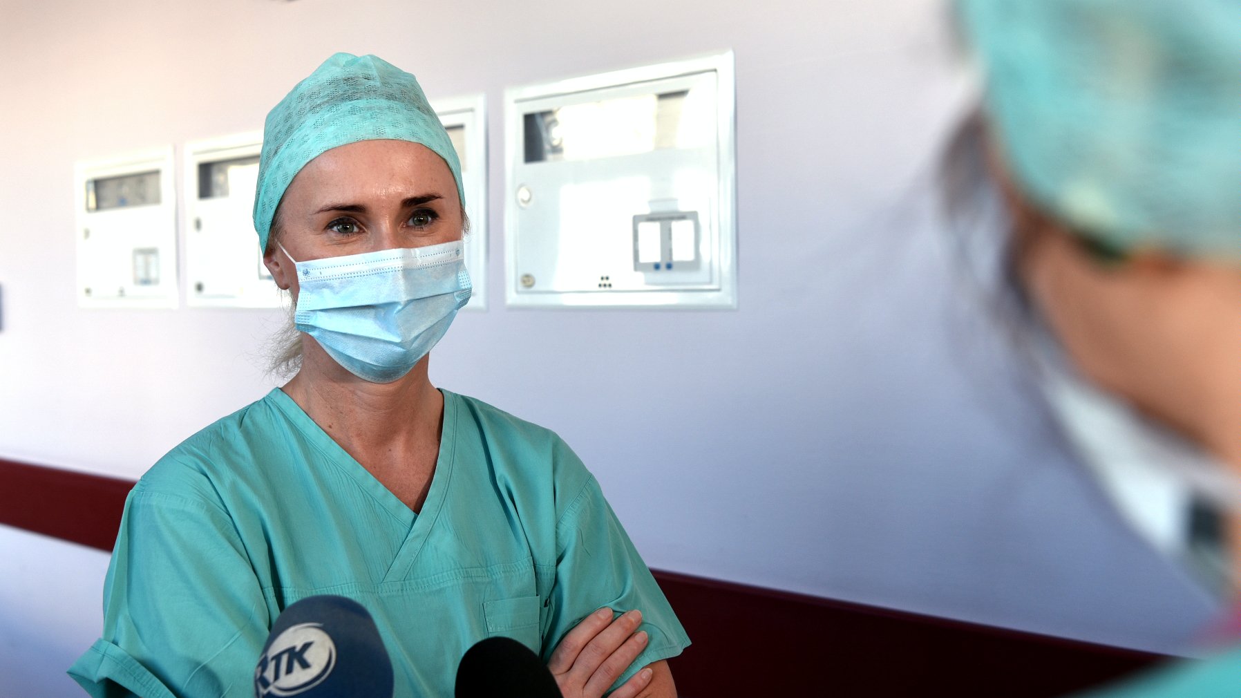 Na zdjęciu lekarka mówiąca coś do mikrofonu, stojąca w szpitalnym korytarzu