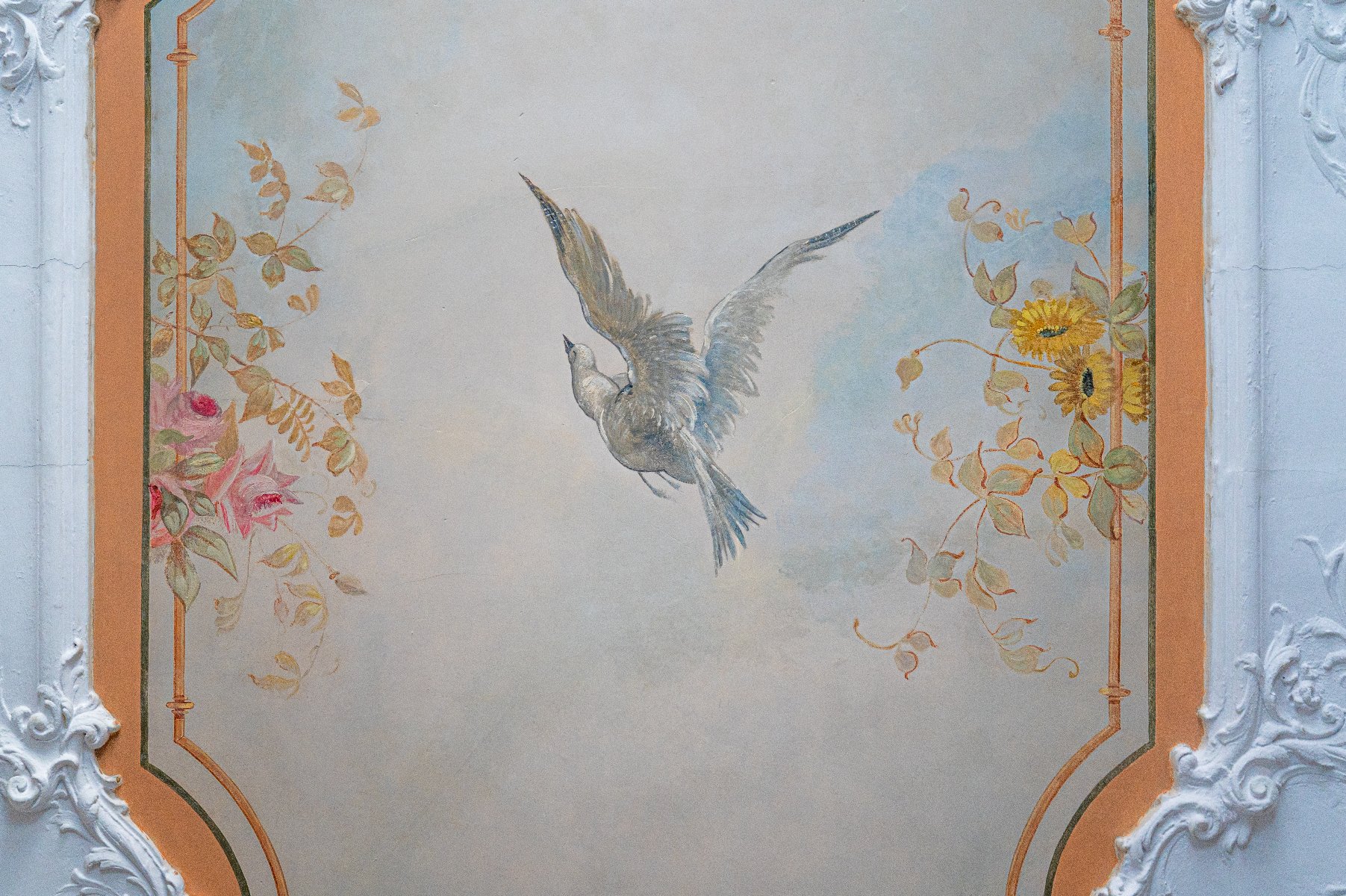 Zbliżenie na dekoracyjny plafon lub fragment ściany w stylu klasycystycznym. W centralnej części, ujętej w ozdobną, cienką ramkę ze złotymi akcentami, namalowany jest lecący ptak na tle jasnego nieba. Po lewej stronie widoczny jest motyw różowych róż, a po prawej żółtych kwiatów przypominających słoneczniki. Całość otacza bogata, biała sztukateria z motywami roślinnymi i zwojami (rolwerkami) na brzoskwiniowym tle.
