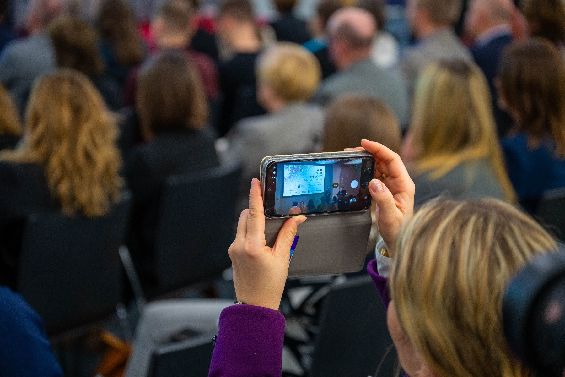 Zbliżenie na dłonie osoby trzymającej smartfon i nagrywającej prezentację podczas konferencji. Na ekranie telefonu widoczny jest slajd wyświetlany na dużym ekranie w głębi sali. W tle rozmyte sylwetki licznie zgromadzonych uczestników siedzących w rzędach.