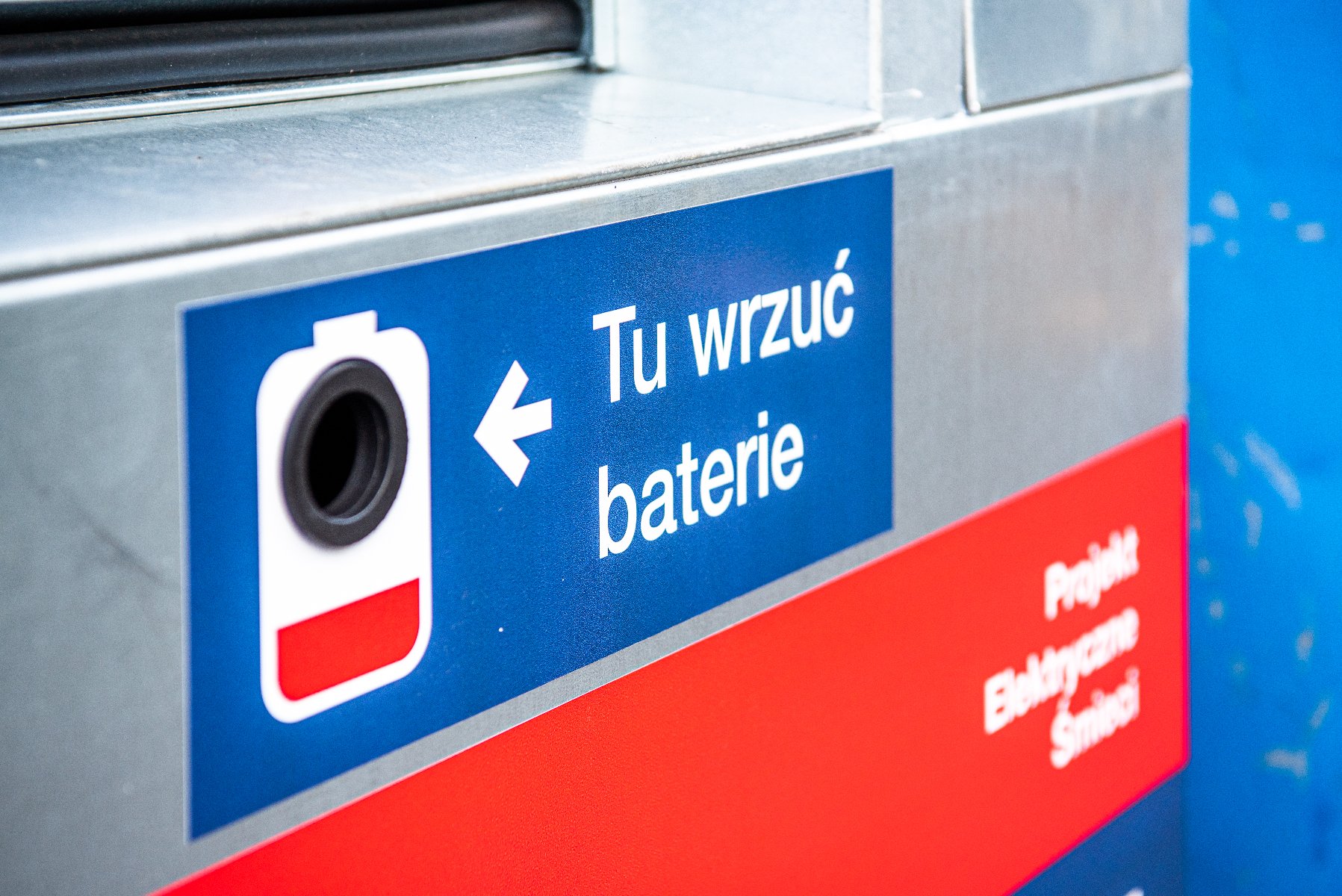 Zbliżenie na otwór wrzutowy pojemnika na odpady elektryczne. Na metalowej obudowie naklejona jest niebieska etykieta z białym napisem »Tu wrzuć baterie«, białą strzałką oraz ikoną baterii. Poniżej znajduje się fragment czerwonej naklejki z napisem »Projekt Elektryczne Śmieci«
