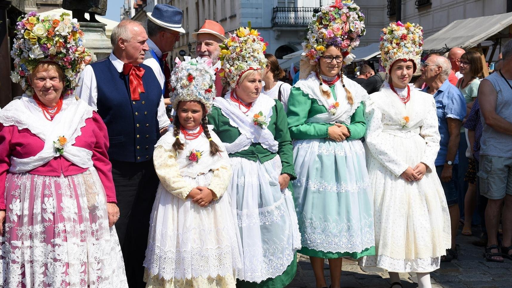 Zdjęcie przedstawia Bambrów Poznańskich na Starym Rynku. Na pierwszym planie widzimy panie przebrane w kolorowe, bogato zdobione suknie z kwiatami na głowie - grafika artykułu