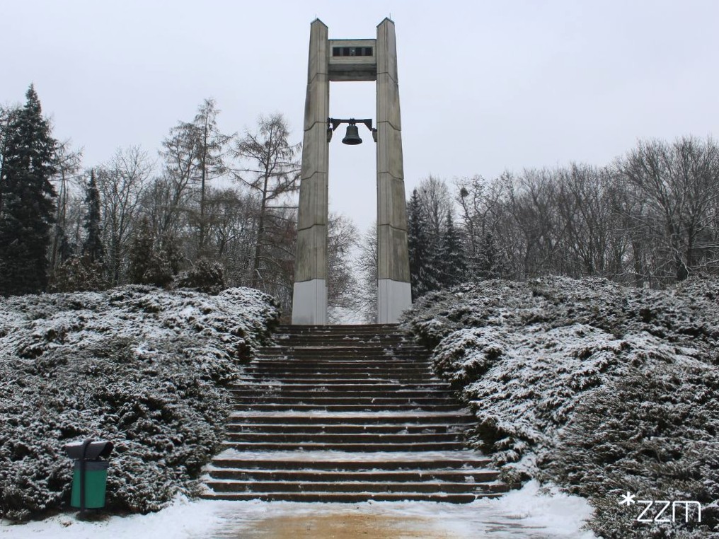 Zdjęcie przedstawia dzwon i pomnik, na którym jest umieszczony oraz schody i zieleń - krzewy i drzewa pokryte śniegiem.