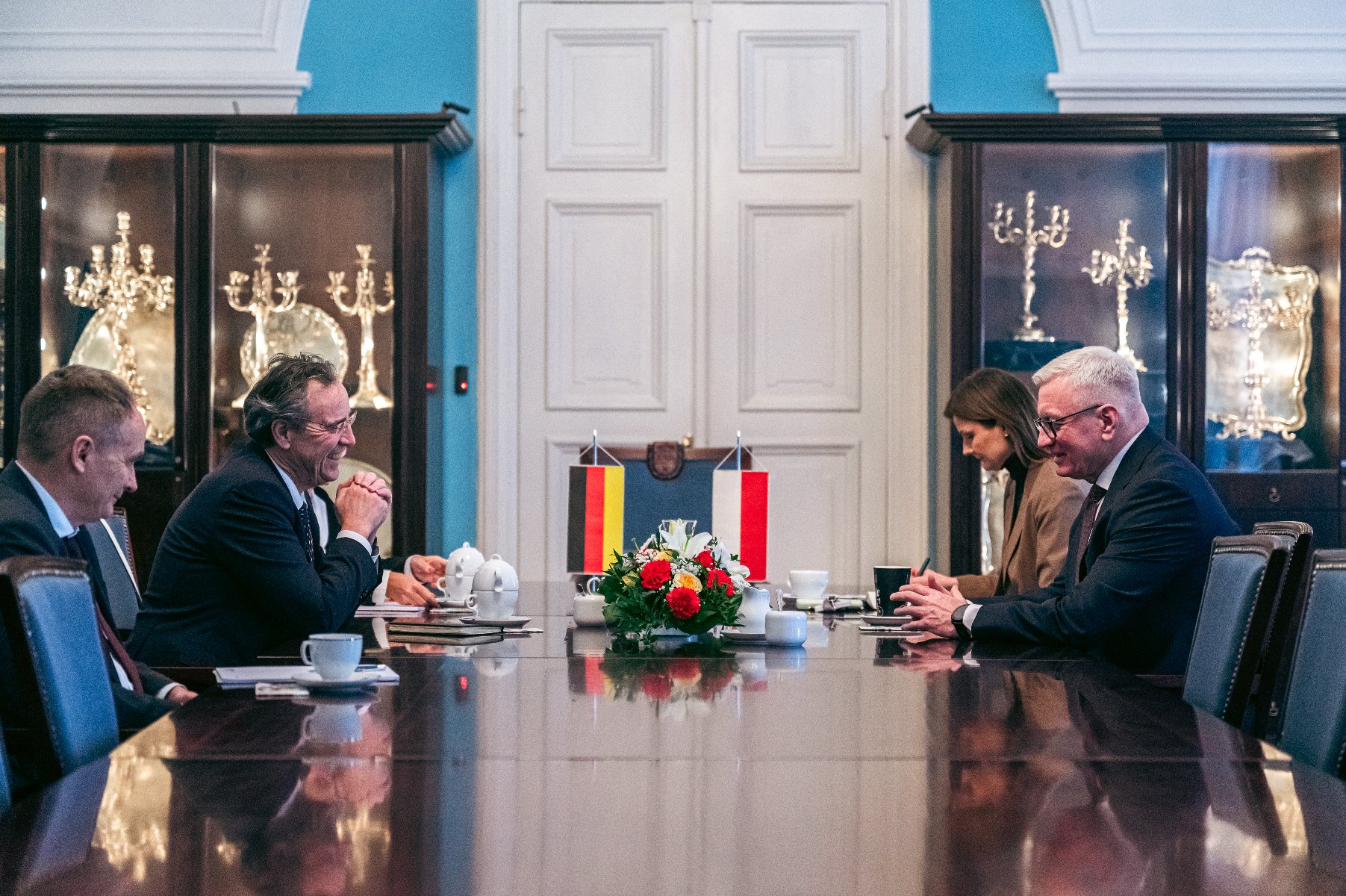 Zdjęcie przedstawia oficjalne spotkanie dwóch delegacji przy długim, lśniącym stole konferencyjnym, na którym ustawiono małe flagi Niemiec i Polski oraz kompozycję z czerwonych i żółtych kwiatów. Po obu stronach stołu siedzą mężczyźni w ciemnych garniturach zaangażowani w rozmowę.
