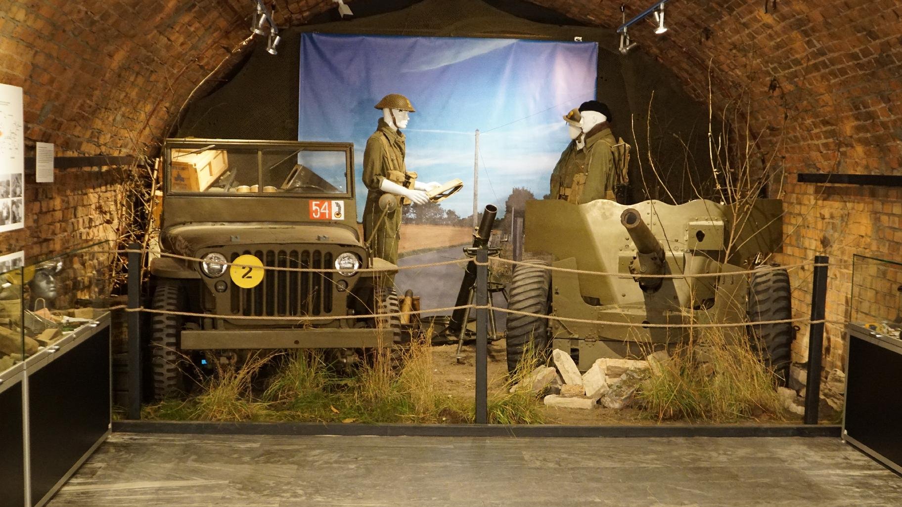 Zdjęcie przedstawia samochód wojskowy i działo. Na drugim planie manekiny ubrane w mudury. - grafika artykułu