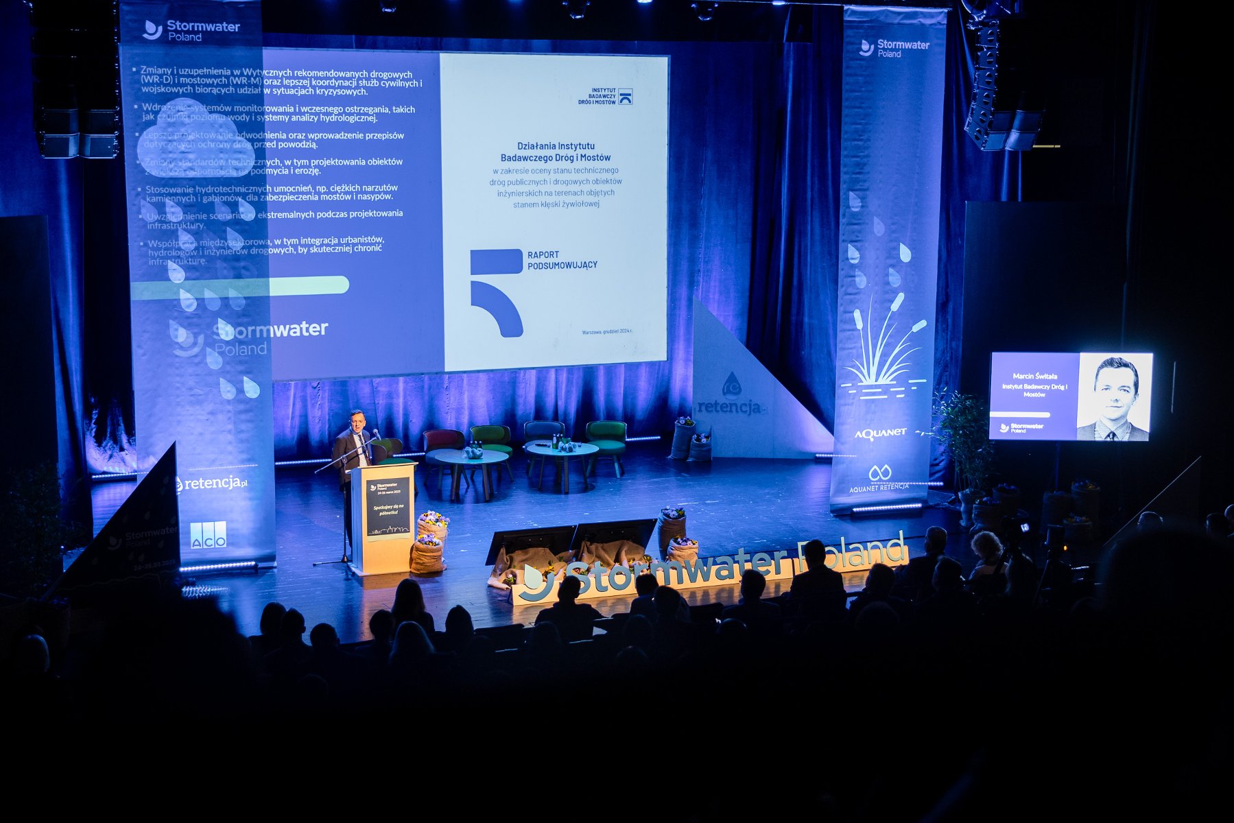 Zdjęcie przedstawia scenę. Przy mównicy stoi kobieta. Za jej plecami wyświetlane są zdjęcia zalanych ulic. Przed nią znajduje się napis Stormwater Poland.
