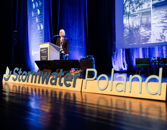 Zdjęcie przedstawia scenę. Przy mównicy stoi kobieta. Za jej plecami wyświetlane są zdjęcia zalanych ulic. Przed nią znajduje się napis Stormwater Poland.