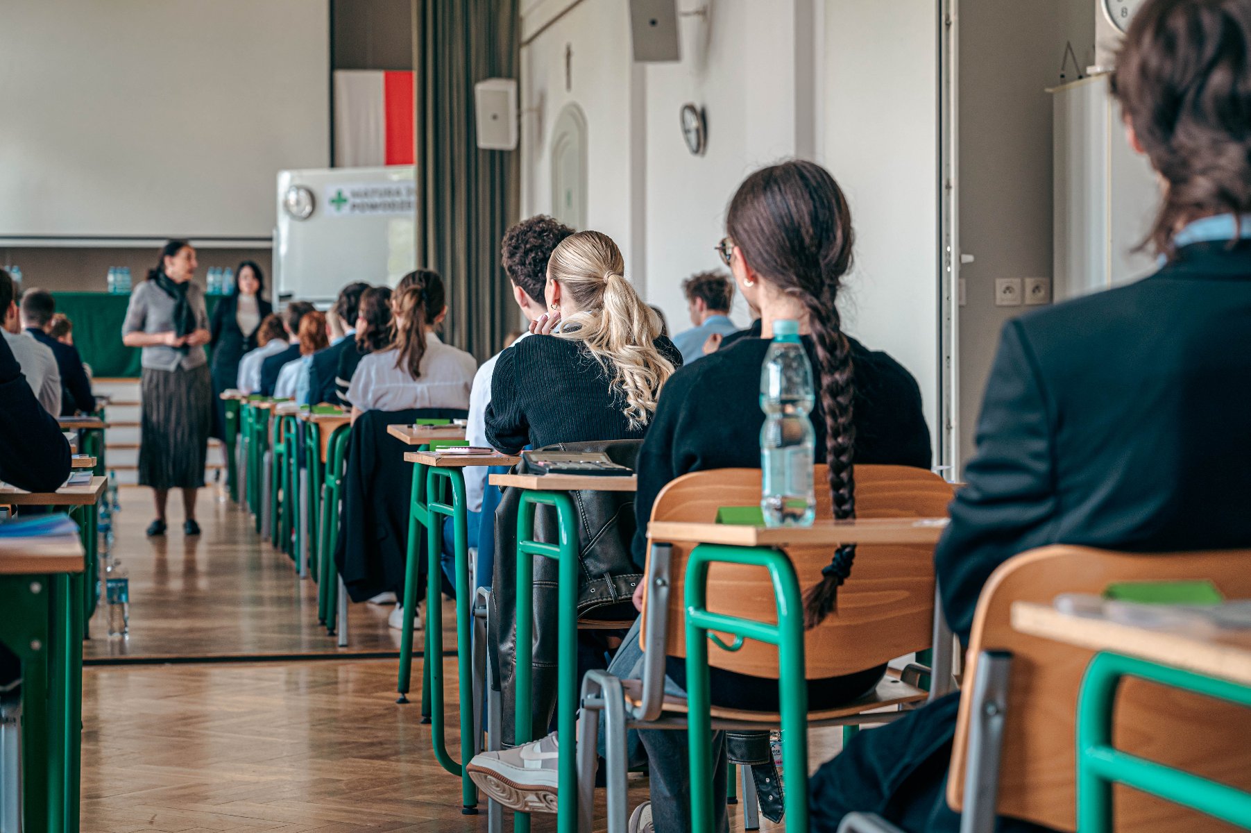 Matura w Poznaniu - ruszyły egzaminy Zdjęcie przedstawia uczniów siedzących w ławkach na sali gimnastycznej, tyłem do aparatu.