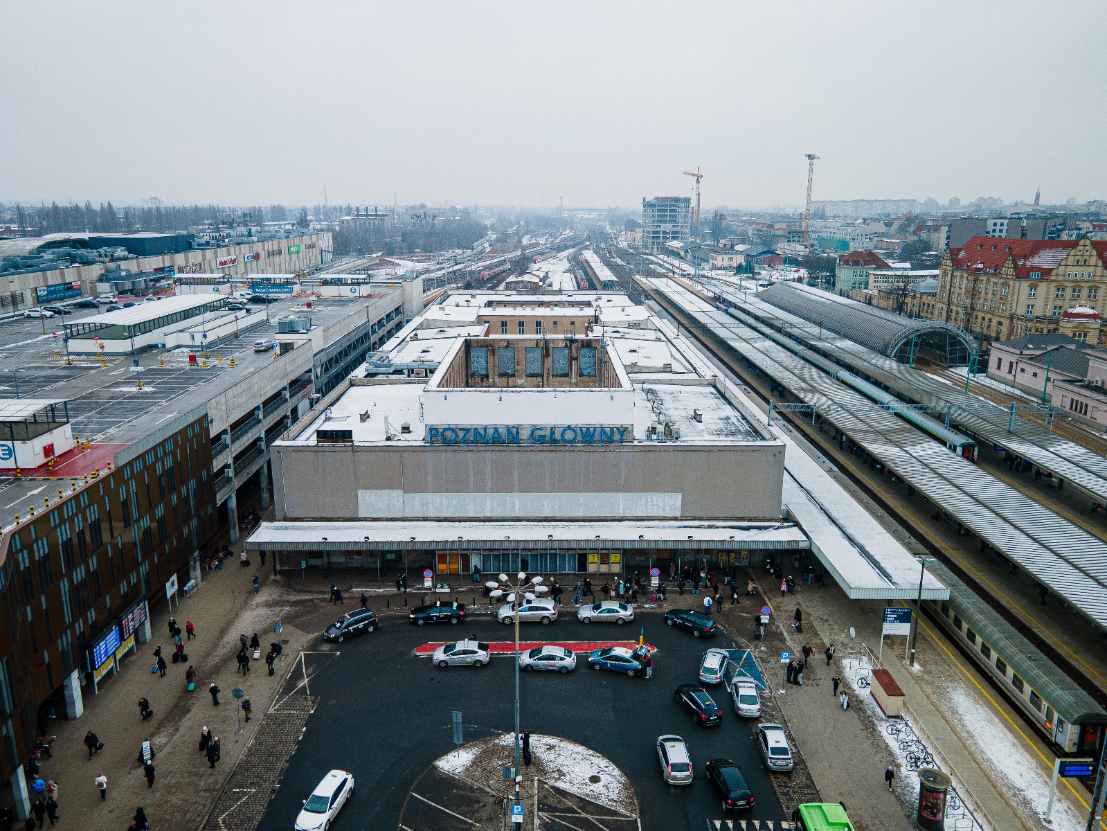 Zdjęcie przedstawia widok z lotu ptaka na stary budynek dworca kolejowego Poznań Główny w zimowej scenerii. Na dachu obiektu, przykrytym cienką warstwą śniegu, widnieje duży niebieski napis z nazwą stacji, a przed wejściem znajduje się półkolisty podjazd, na którym parkują liczne taksówki.