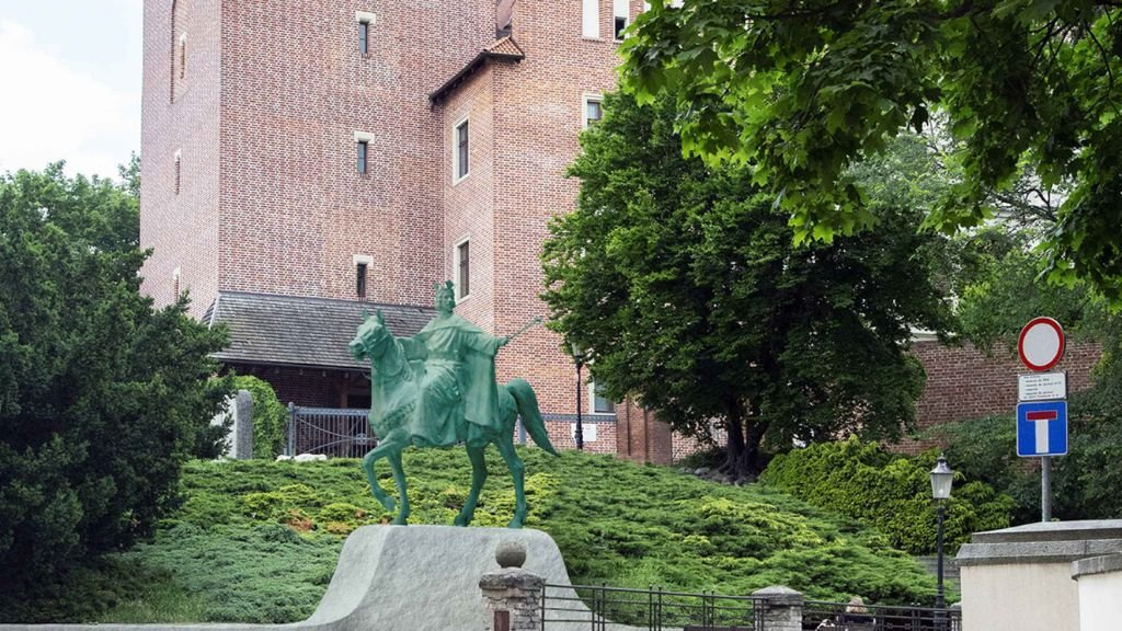 Zielony, metalowy pomnik na koniu stojący na szarym, kamiennym postumencie. Za pomnikiem wznosi się duża, ceglana ściana zabytkowego budynku z małymi oknami. Po prawej stronie widoczne drzewa i znak drogowy.