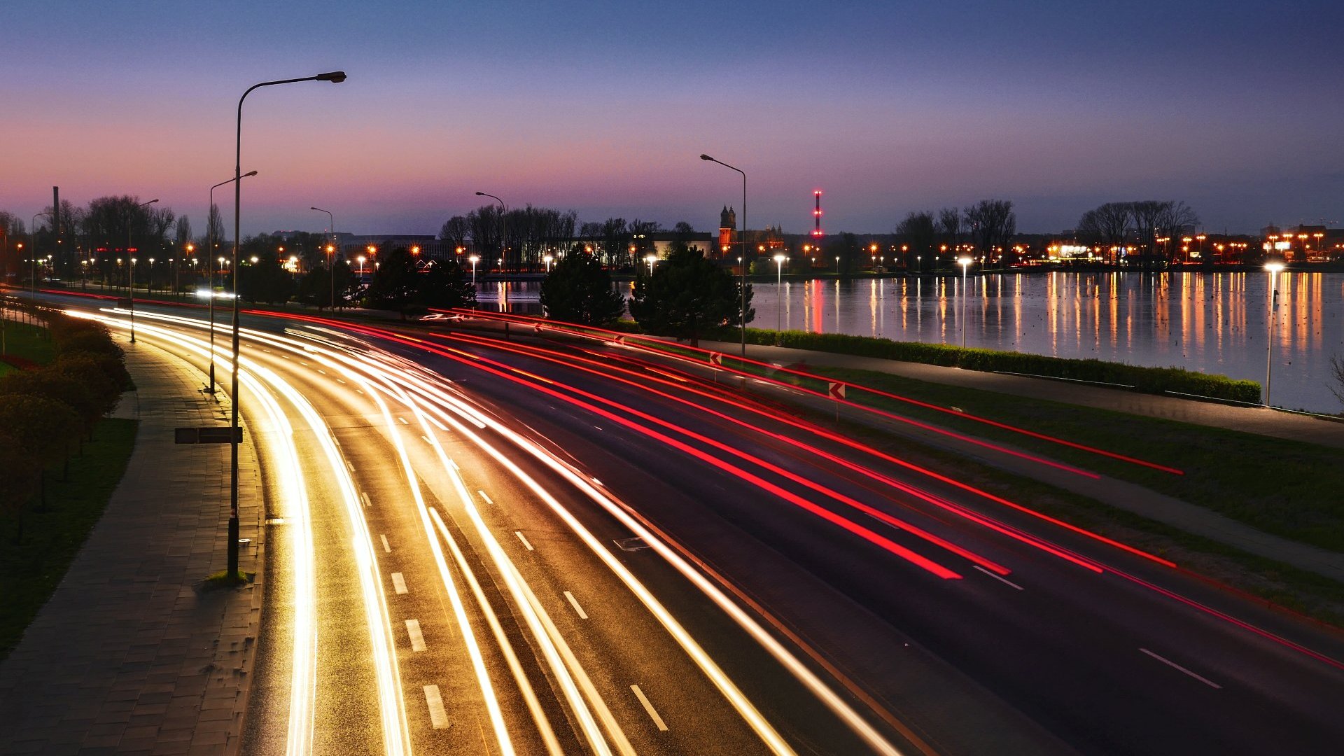 a photo of a street illuminated by lights. next to it there's the Malta Lake and the Poznań Cathedral in the background
