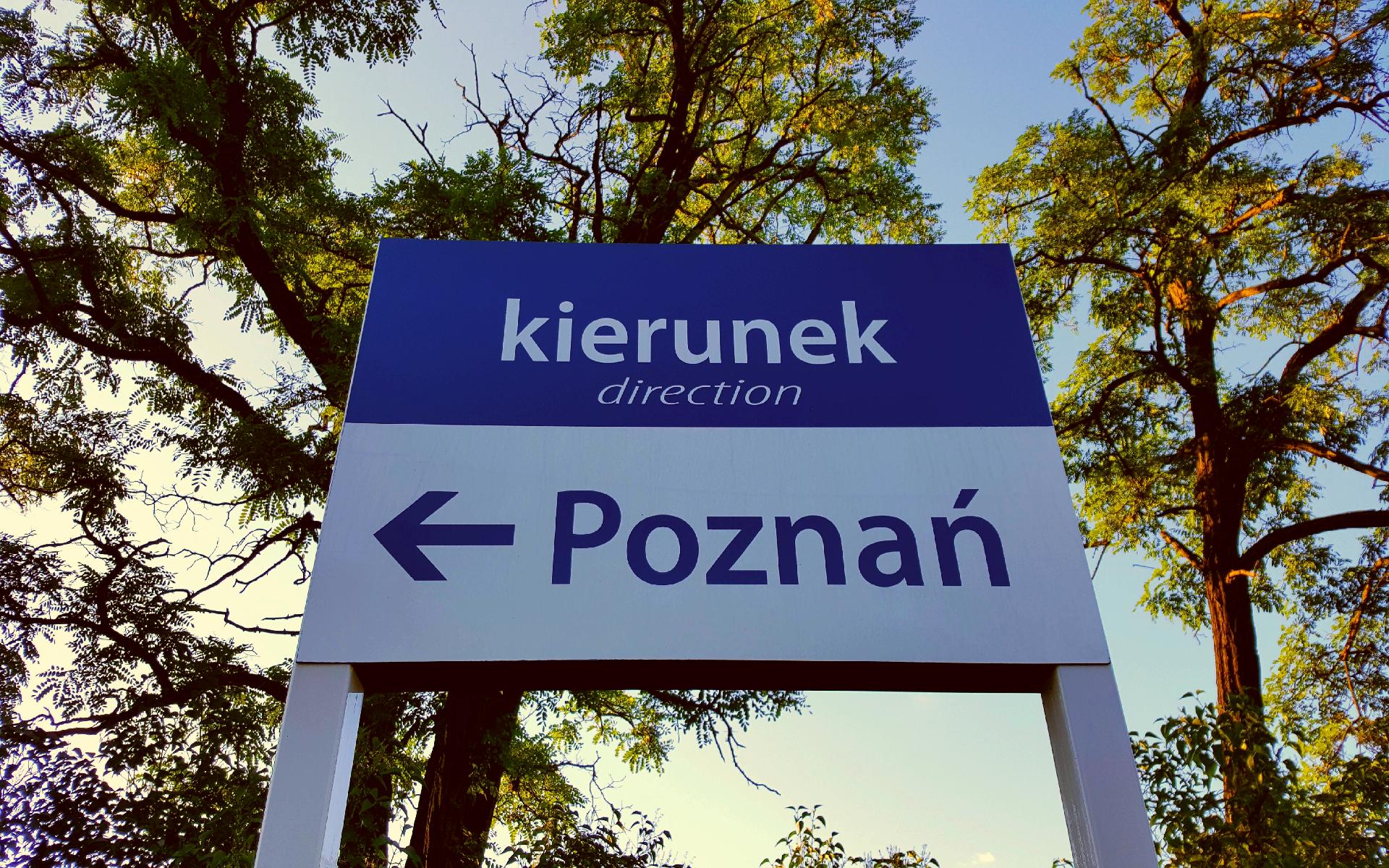 a sign on a train station that says: direction: Poznań. trees in the background
