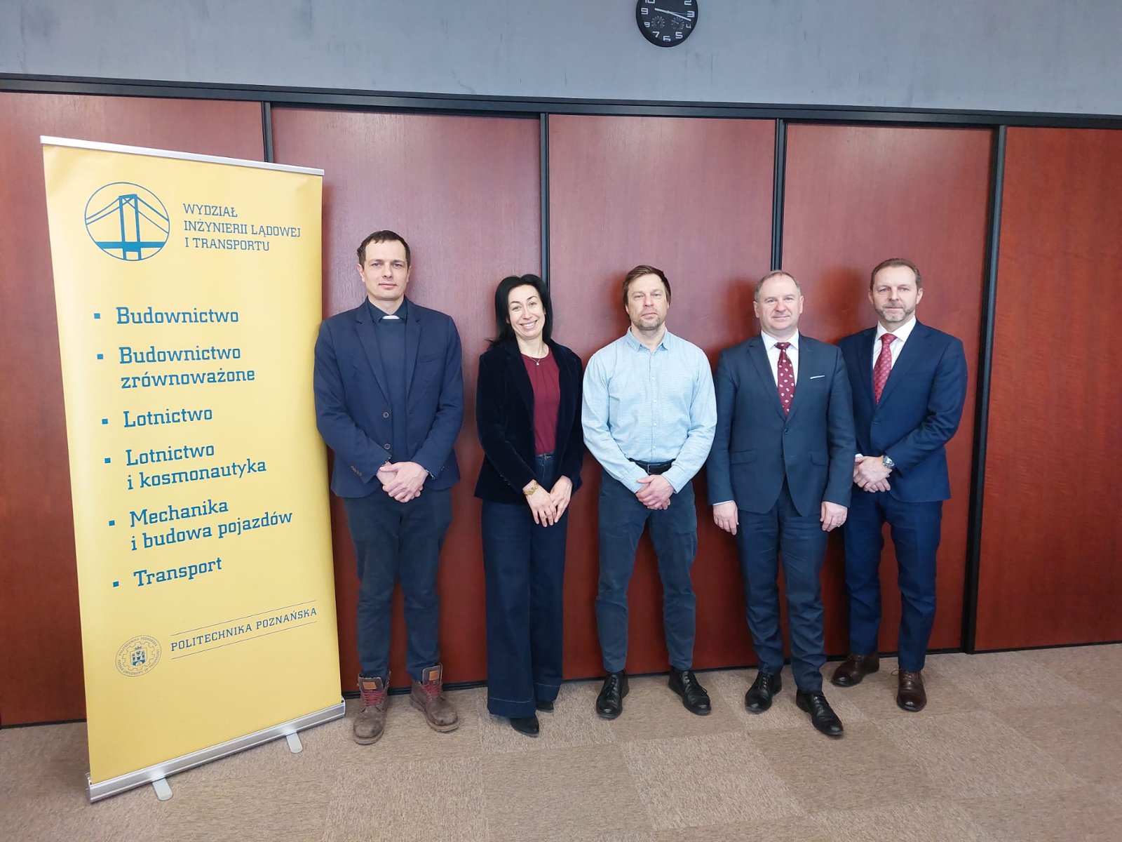 Five people (four men and one woman) standing in a row in front of a brown wall. To the left is a yellow roll-up banner of the Faculty of Civil and Transport Engineering of Poznan University of Technology, listing study programs such as Civil Engineering, Aviation, and Transport
