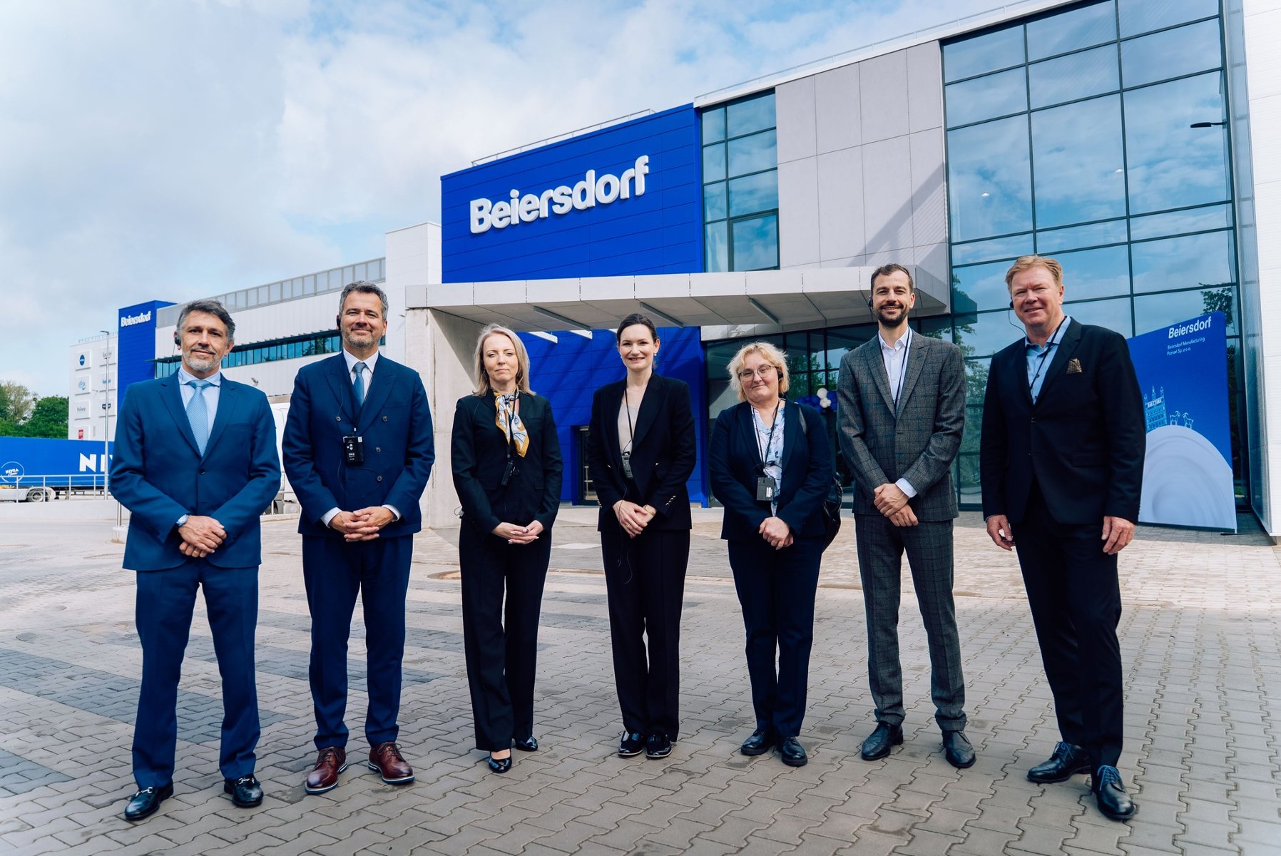 Seven people in business attire stand before a modern Beiersdorf facility. The group consists of four men and three women, some wearing communication headsets. The building features a large blue facade with white branding. To the left, a Nivea truck is visible, highlighting the company's skincare portfolio. The scene is set on a paved courtyard under a bright, slightly cloudy sky, suggesting a corporate event or factory visit.