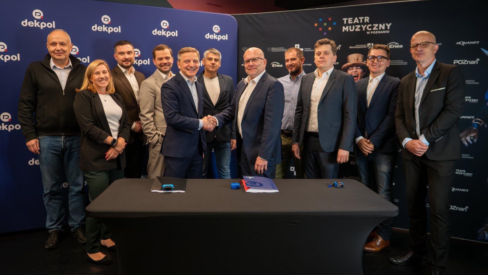 An agreement has been signed for the construction of the new headquarters of the Musical Theatre in Poznań Two men shaking hands. In the background, a group of elegantly dressed people. They are celebrating the signing of the agreement.