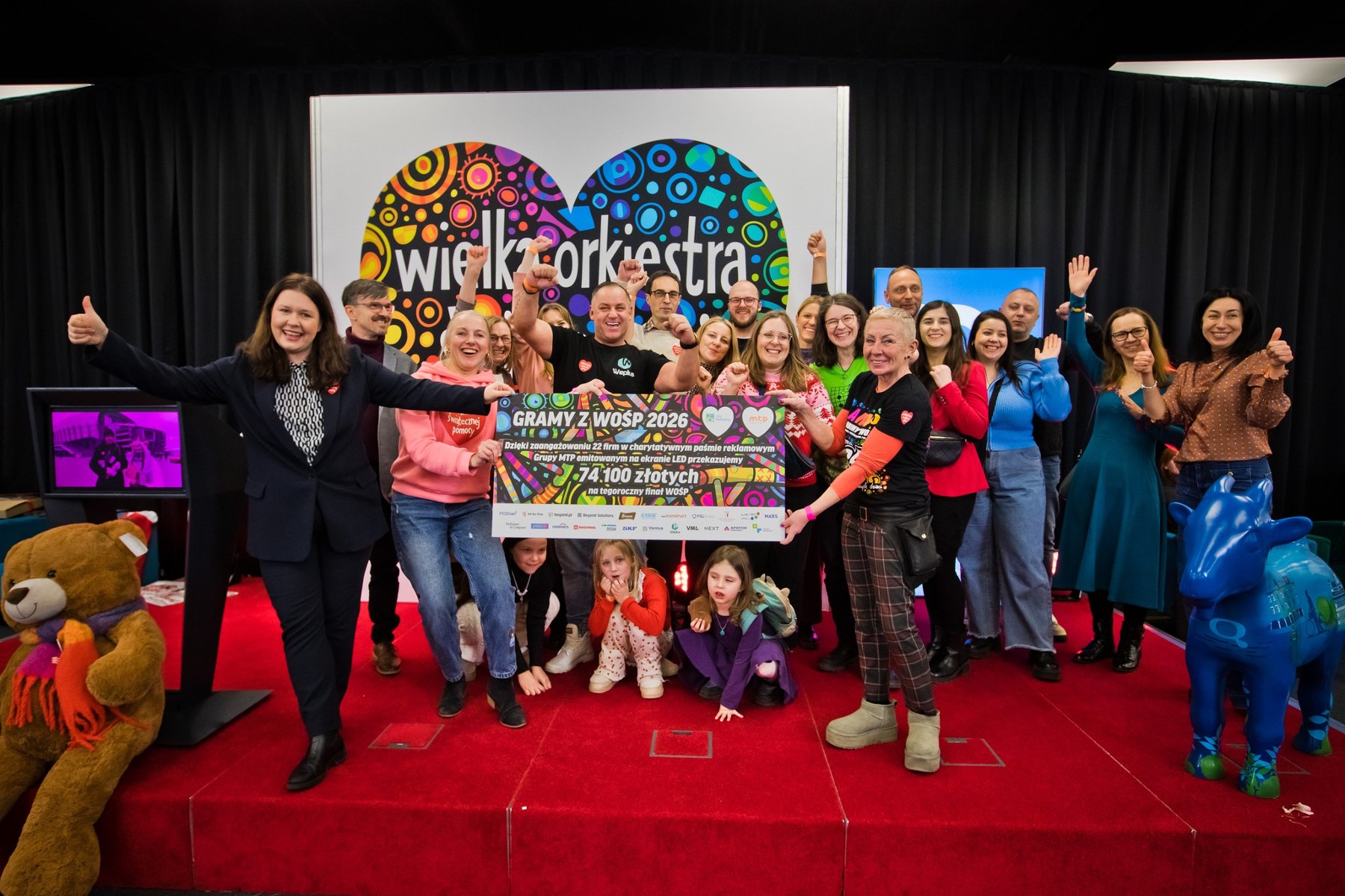 Grupa uśmiechniętych dorosłych i dzieci stoi na czerwonym podeście, trzymając wielki, pamiątkowy czek na kwotę 74 100 złotych dla Wielkiej Orkiestry Świątecznej Pomocy. W tle widać duże, kolorowe logo WOŚP w kształcie serca, a po bokach sceny dużego pluszowego misia oraz niebieską figurkę krowy.