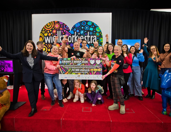 Grupa uśmiechniętych dorosłych i dzieci stoi na czerwonym podeście, trzymając wielki, pamiątkowy czek na kwotę 74 100 złotych dla Wielkiej Orkiestry Świątecznej Pomocy. W tle widać duże, kolorowe logo WOŚP w kształcie serca, a po bokach sceny dużego pluszowego misia oraz niebieską figurkę krowy.