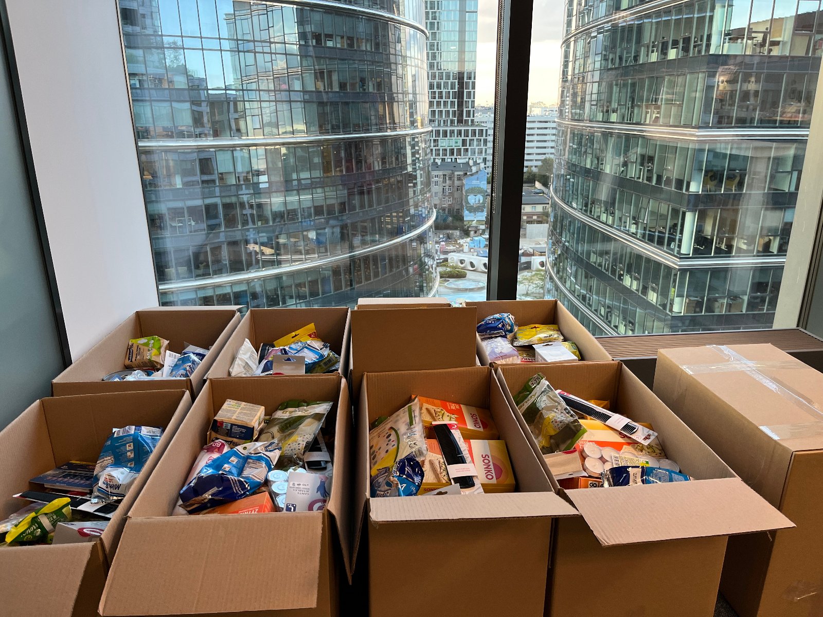 Kartony z żywnością i produktami higieny dla powodzian. Ułożone w kartonach leżą pod oknem, w tle widać biurowce.