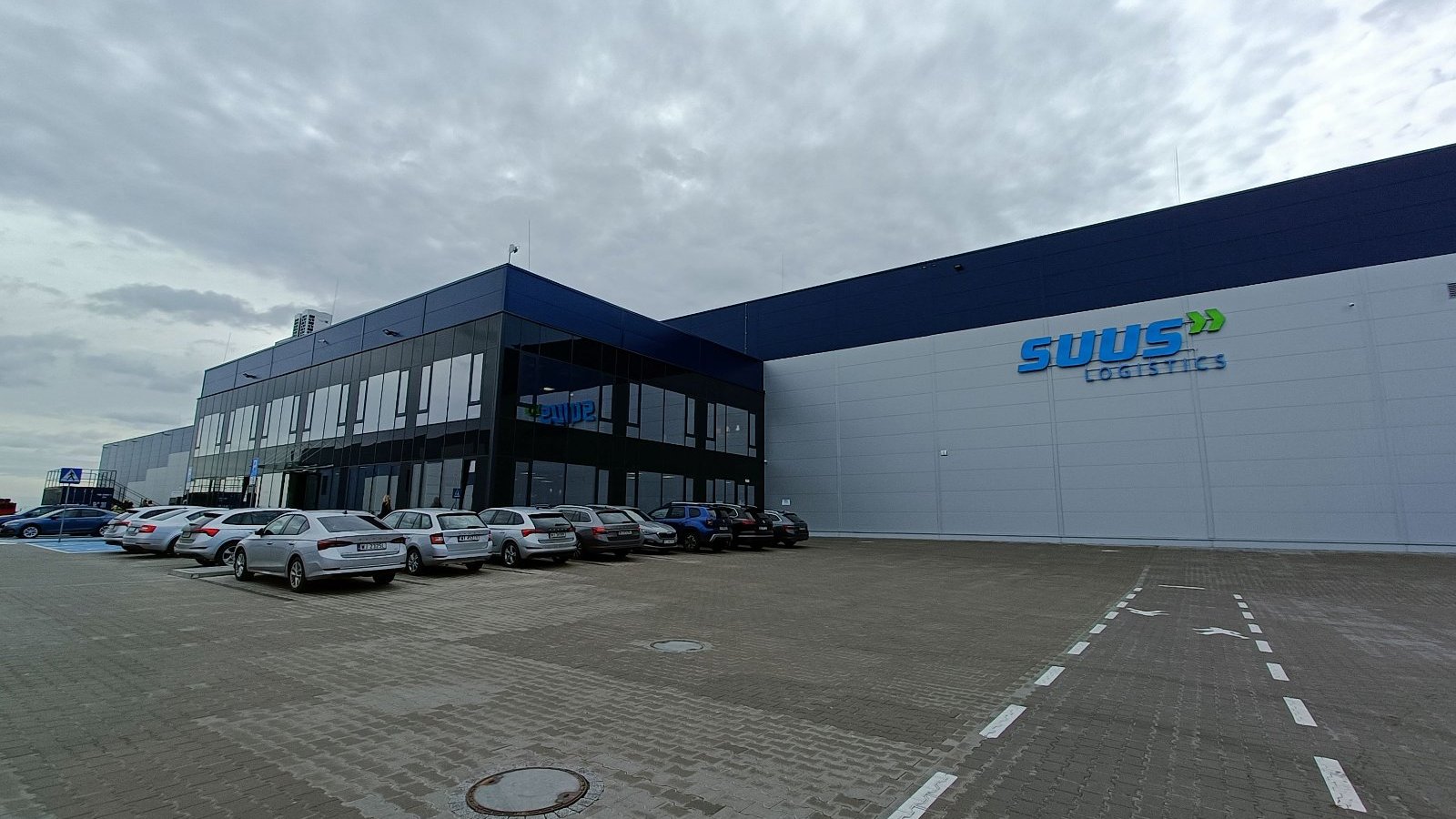 Zdjęcie przedstawia budynek magazynowy firmy SUUS Logistics. Przed magazynem znajduje się szklane wejście. Na parkingu stoją samochody osobowe.