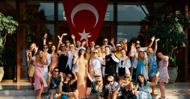 Zdjęcie przedstawia pracowniczki i pracowników firmy pozujących do wspólnego zdjęcia. W tle znajduje się przeszklony budynek z flagą Turcji.
