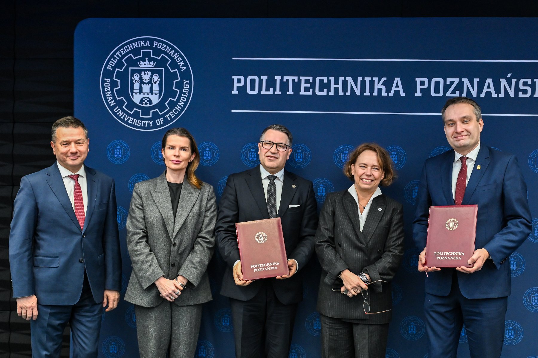 Pięć osób w biznesowych strojach (trzech mężczyzn i dwie kobiety) stoi na tle ciemnoniebieskiej ściany z logotypem i napisem "POLITECHNIKA POZNAŃSKA". Mężczyzna w środku i mężczyzna po prawej trzymają w dłoniach dokumenty oprawione w bordową skórę.