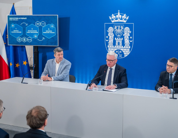 Prezydent Poznania w trakcie konferencji prasowej. W tle ściana z herbem Poznania.