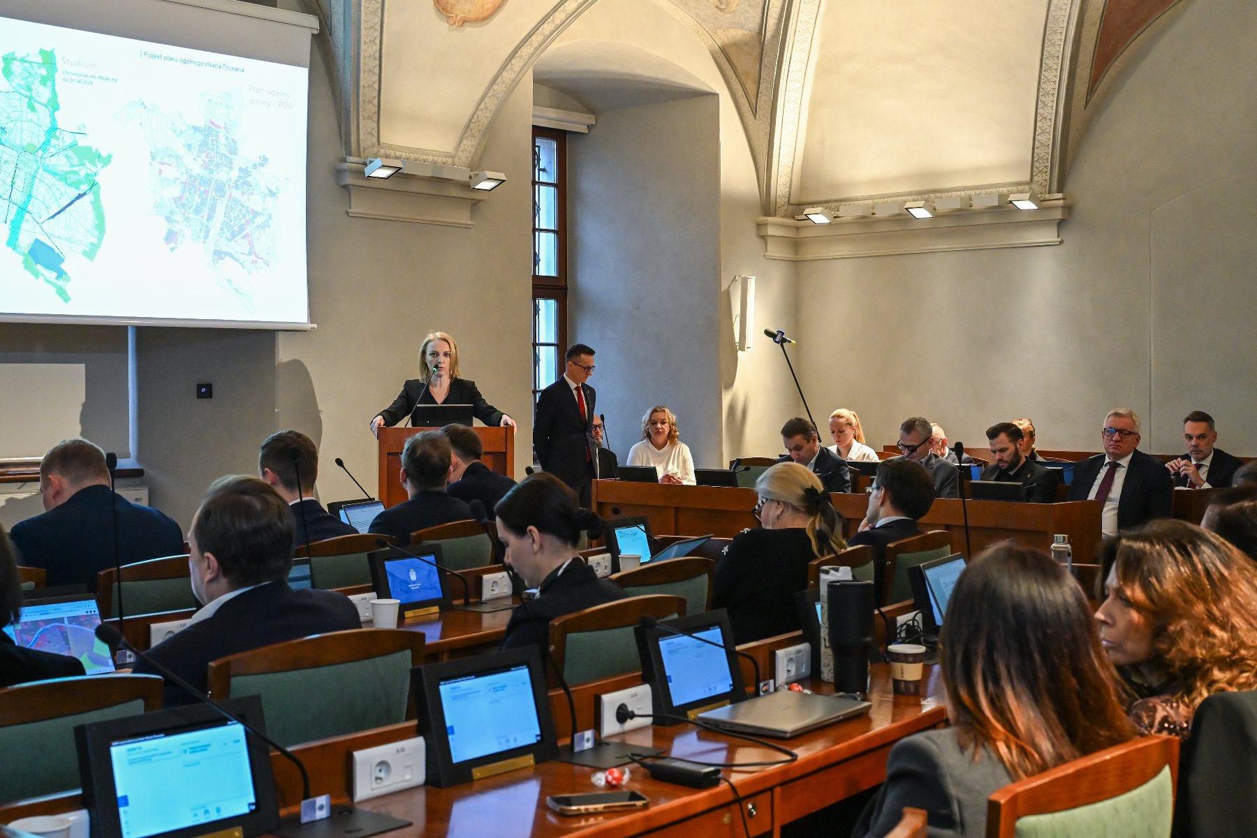 Zdjęcie z sali obrad, przedstawiające liczną grupę osób siedzących przy długich stołach wyposażonych w małe ekrany monitorów. W centrum, przy drewnianej mównicy, stoi kobieta w czarnej marynarce i przemawia do mikrofonu.