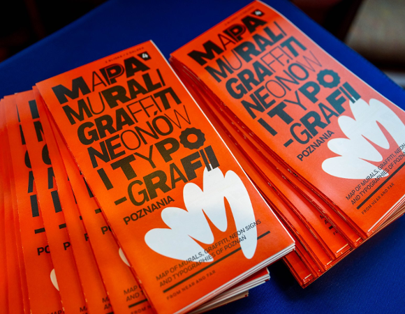 Dwa stosy pomarańczowych ulotek lub broszur rozłożone na niebieskim podłożu. Na okładkach widnieje duży, czarny napis w języku polskim: »MAPA MURALI, GRAFFITI, NEONÓW I TYPOGRAFII POZNANIA«. Poniżej znajduje się biała grafika przypominająca dłoń lub kwiat oraz mniejszy napis w języku angielskim.