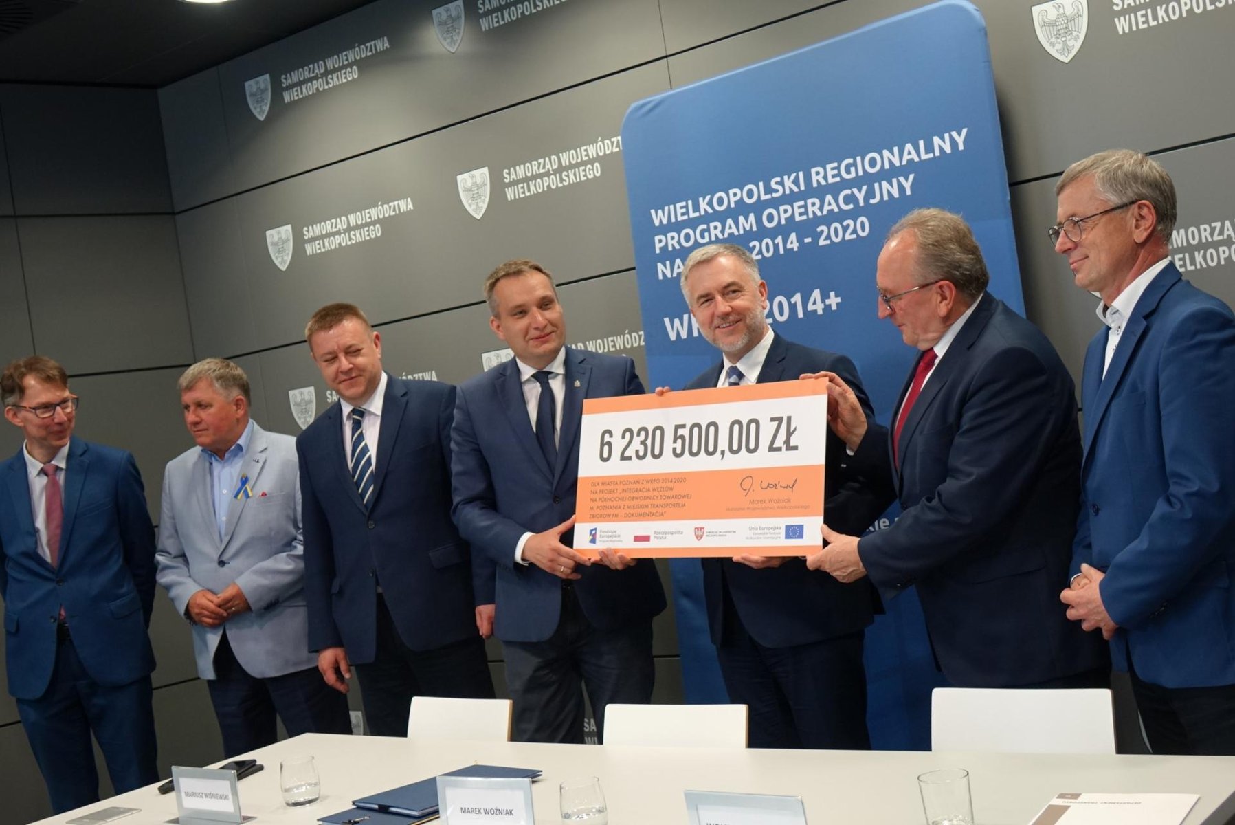 Na zdjęciu grupa mężczyzn w garniturach trzymająca planszę z "czekiem" - grafika artykułu