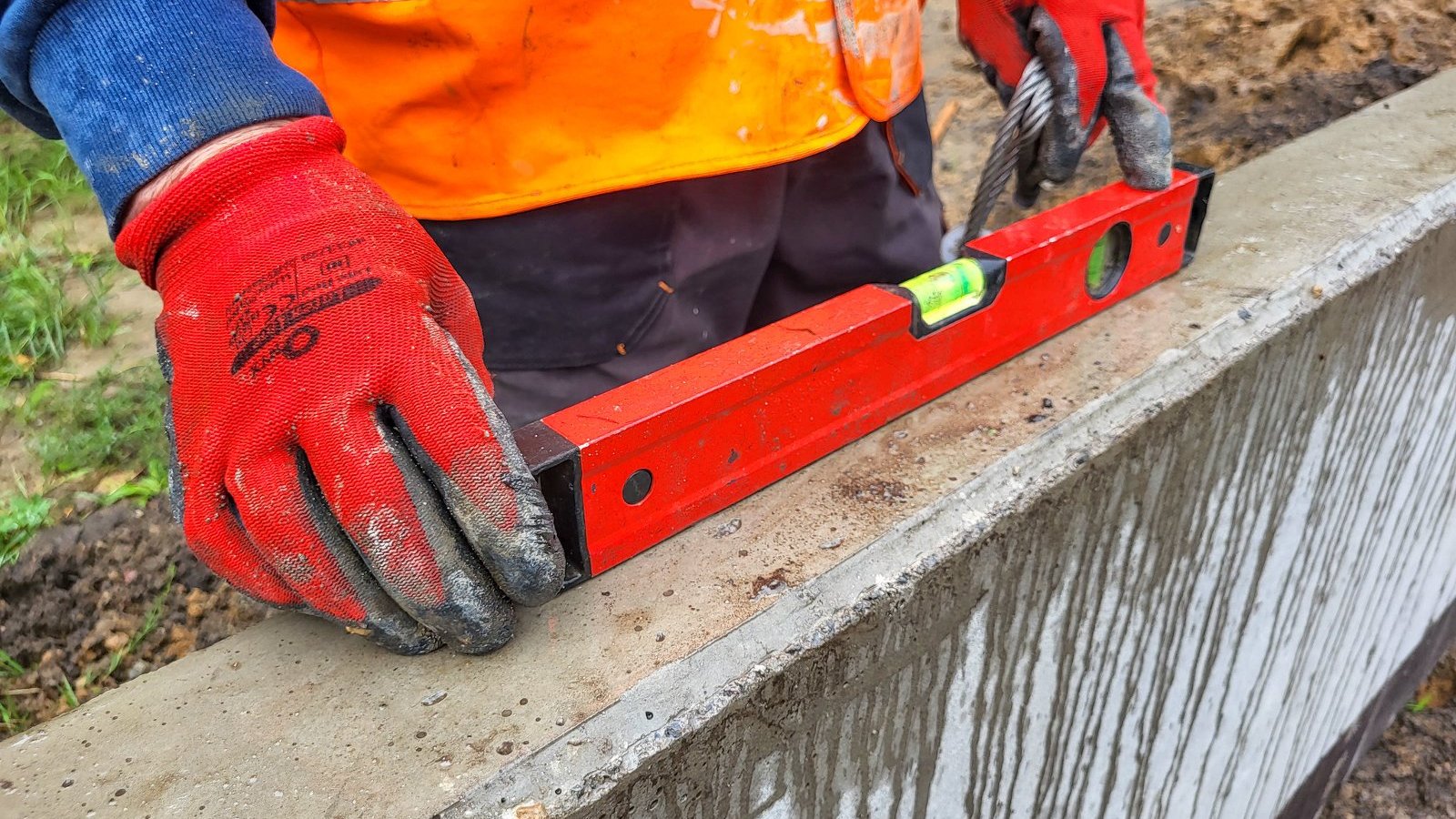Zdjęcie przedstawia ręcę mężczyzny w rękawiczkach i roboczym ubraniu, który sprawdza poziomicą, czy mur jest równy.
