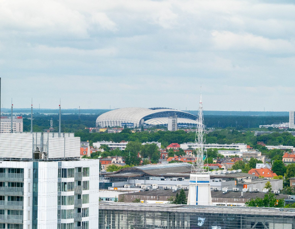 Panorama Poznania z widokiem na Stadion Miejski na horyzoncie. Na pierwszym planie widać nowoczesne budynki mieszkalne i biurowe. Wokół stadionu widać drzewa i zieleń, a w tle rozciąga się las.