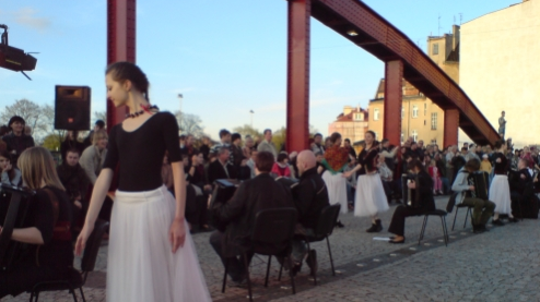 Na fotografii widać publiczność siedzącą przys tolikach, które znajduja sie na Moście nad Cybiną. Na pierwszym planie tancerka ubrana w białą spódnicę i czarną bluzkę.