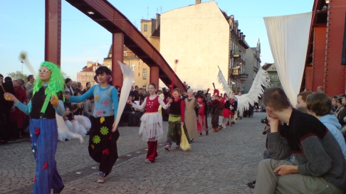 Na zdjęciu widać korowód przbranej młodzieży za kolorowe postacie. Korowód przechodzi mostem. W tle kamienice.