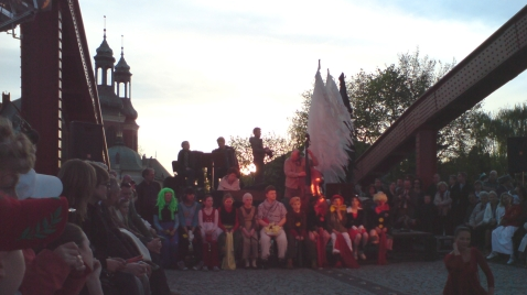 Młodzież siedząca na moście. Po bokach czerwone przęsła mostu. W tle katedra.